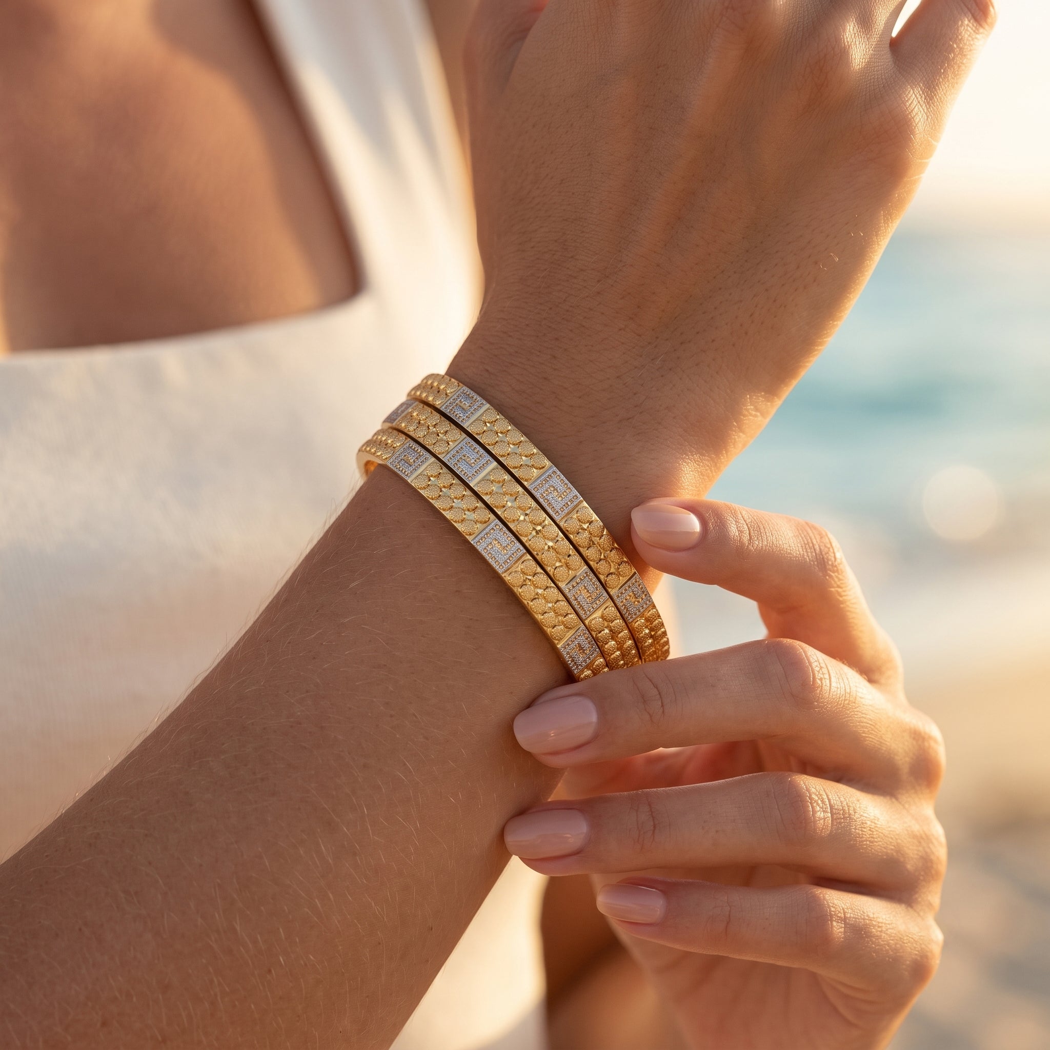Extreme macro close-up of three perfectly stacked E.Z two-tone patterned gold bangles on a woman's wrist during golden hour.	لقطة مقربة جداً لثلاث غوايش منقوشة بلونين متناسقة من E.Z على معصم امرأة وقت الغروب.