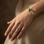 Hand wearing a gold ring with green gemstones on a brown fabric background