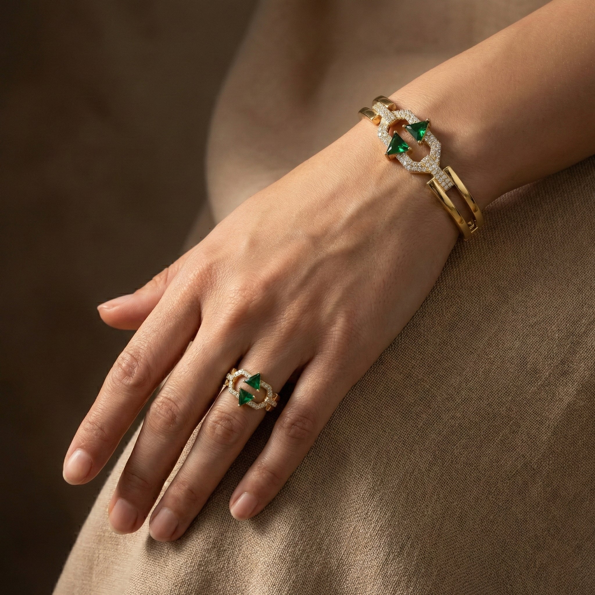 Hand wearing a gold ring with green gemstones on a brown fabric background