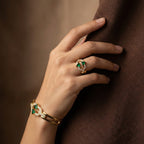 Hand wearing a gold ring with green gemstones on a brown background