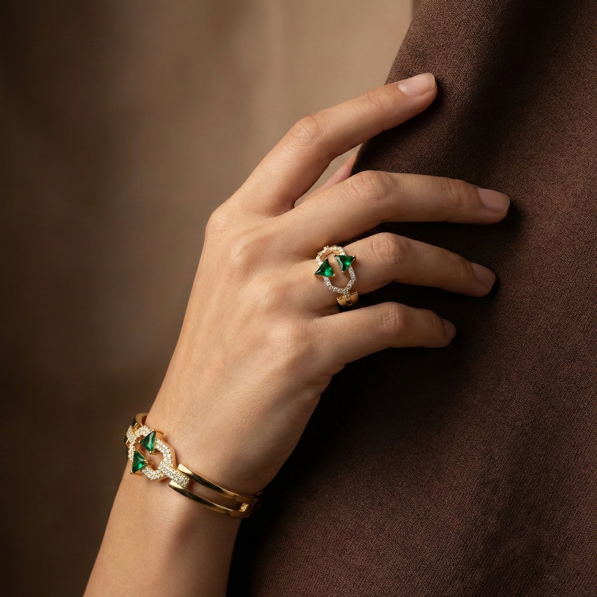 Hand wearing a gold ring with green gemstones on a brown background