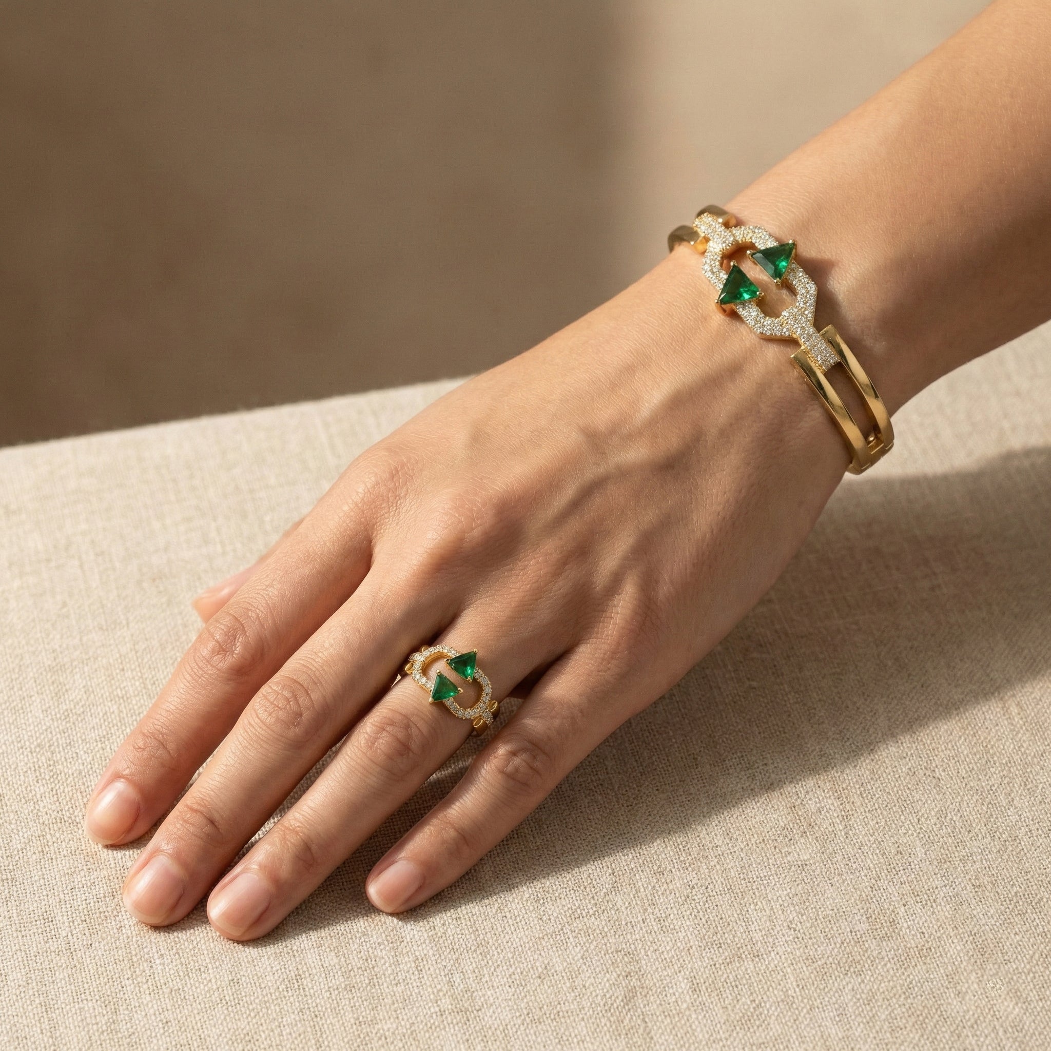 Hand wearing a gold bracelet and ring with green gemstones on a beige surface