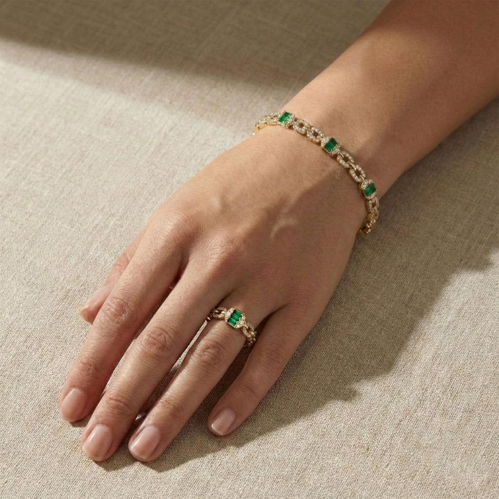 Hand wearing a gold bracelet and ring with green gemstones on a beige fabric background