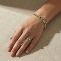 Hand wearing a gold bracelet and ring with green gemstones on a beige fabric background