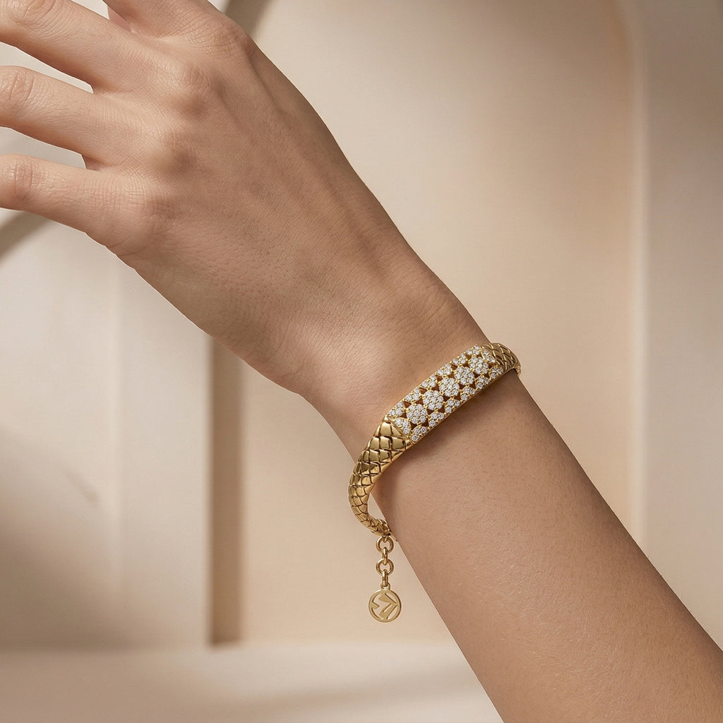 Editorial photograph of a Lazurde 18k yellow gold woven bracelet on a feminine wrist. The hand is posed sculpturally against a minimalist nude-beige background with soft architectural arches and delicate shadow play.