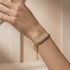 Editorial photograph of a Lazurde 18k yellow gold woven bracelet on a feminine wrist. The hand is posed sculpturally against a minimalist nude-beige background with soft architectural arches and delicate shadow play.