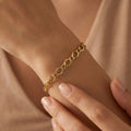 An ultra-realistic close-up of the 3B 18k yellow gold Gourmet bracelet worn on a feminine wrist, with the other hand gently touching it against a soft nude background in warm diffused lighting.