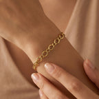 An ultra-realistic close-up of the 3B 18k yellow gold Gourmet bracelet worn on a feminine wrist, with the other hand gently touching it against a soft nude background in warm diffused lighting.