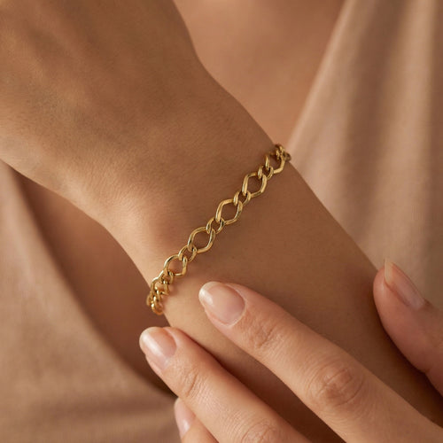An ultra-realistic close-up of the 3B 18k yellow gold Gourmet bracelet worn on a feminine wrist, with the other hand gently touching it against a soft nude background in warm diffused lighting.