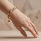 An elegant editorial photo of a Lazurde 18k yellow gold bracelet styled on a feminine wrist. The hand is posed sculpturally, resting lightly on a surface against a warm beige background with subtle abstract shapes, emphasizing the polished gold links and intricate center detail.