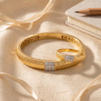 A pair of gold bangles with textured bands and square diamond-set accents, arranged on cream satin fabric with a pencil and notebook in the background.