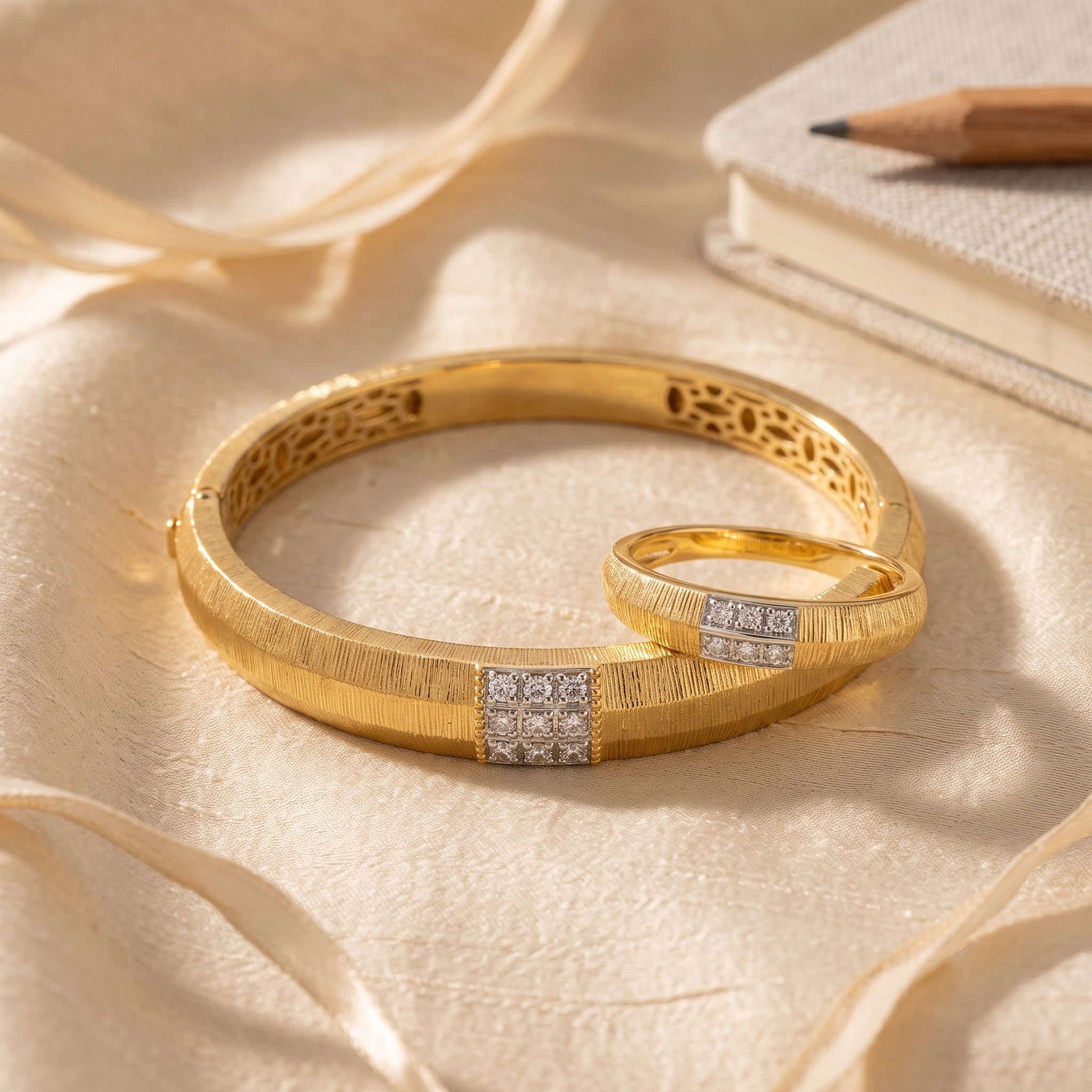 A pair of gold bangles with textured bands and square diamond-set accents, arranged on cream satin fabric with a pencil and notebook in the background.