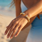 Extreme close-up of a woman's gracefully posed wrist wearing a UNO gold open cuff bracelet against a blurred beach background.	لقطة مقربة جداً لمعصم امرأة ترتدي إسورة أونو الذهبية المفتوحة بأناقة، مع خلفية شاطئ مموهة.