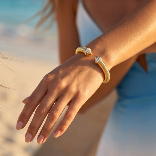 Extreme close-up of a woman's gracefully posed wrist wearing a UNO gold open cuff bracelet against a blurred beach background.	لقطة مقربة جداً لمعصم امرأة ترتدي إسورة أونو الذهبية المفتوحة بأناقة، مع خلفية شاطئ مموهة.