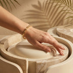 Hand wearing the L'azurde 18k gold bracelet resting elegantly on a warm ivory sculptural pedestal with tropical palm shadows.	يد ترتدي انسيال ذهب لازوردي عيار 18 تستريح بأناقة على منصة منحوتة بلون عاجي دافئ مع ظلال استوائية لأوراق النخيل.