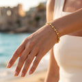 Feminine wrist wearing the delicate L'azurde 18k gold square link bracelet against a blurred Mediterranean beach background.	معصم أنثوي يرتدي انسيال ذهب لازوردي الرقيق عيار 18 بحلقات مربعة مع خلفية ضبابية لشاطئ متوسطي.