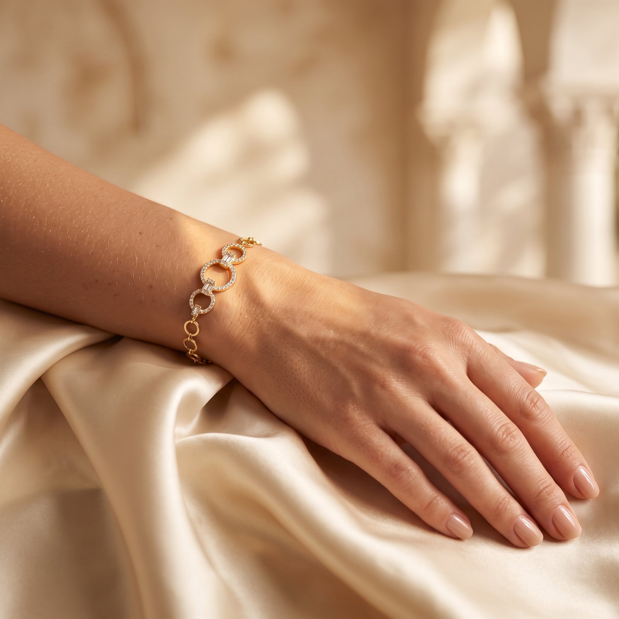 A close-up view of a woman's forearm resting on cream satin, wearing a L'azurde gold bracelet (5.12g) with interlocking stone-paved links. Soft light and palm shadows are in the background. لقطة قريبة لساعد امرأة يستريح على ساتان بلون الكريم، ترتدي سواراً ذهبياً من لازوردي (بوزن 5.12 غرام) يتميز بحلقات متشابكة مرصعة بالأحجار اللامعة. تظهر إضاءة ناعمة وظلال سعف النخيل في الخلفية.