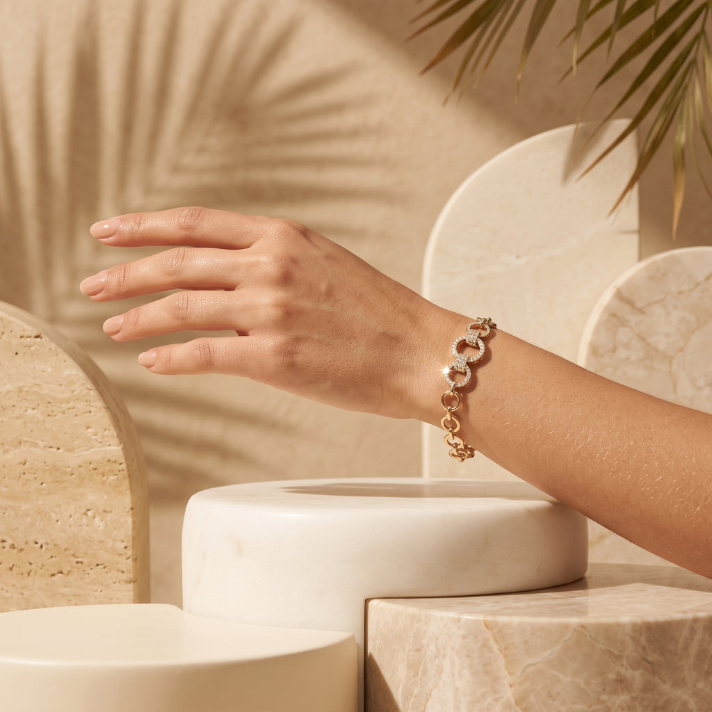 A stylized close-up of a woman's hand wearing a L'azurde gold bracelet (5.12g) on her wrist, set against stacked stone and marble pedestals. Warm sunlight casts textured palm leaf shadows on the back wall. لقطة قريبة فنية ليد امرأة ترتدي سواراً ذهبياً من لازوردي (بوزن 5.12 غرام) على معصمها، معروضة على خلفية من ركائز حجرية ورخامية مكدسة. تسلط أشعة الشمس الدافئة ظلالاً بارزة لسعف النخيل على الجدار الخلفي.