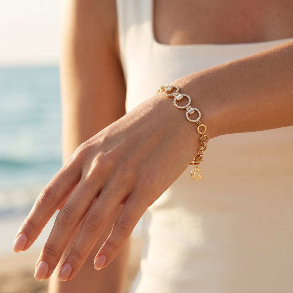 A close-up of a woman's hand wearing a L'azurde gold bracelet (5.12g) with stone-paved interlocking links, set against a sun-drenched beach and ocean. Fingers are slightly curved, and the brand charm dangles from the wrist. لقطة قريبة ليد امرأة ترتدي سواراً ذهبياً من لازوردي (بوزن 5.12 غرام) المزود بحلقات متداخلة مرصعة بالأحجار، معروضة على خلفية من شاطئ وبحر مغمورين بأشعة الشمس. الأصابع منحنية قليلاً، وتتدلى دلاية العلامة التجارية من المعصم.