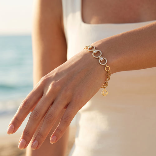 A close-up of a woman's hand wearing a L'azurde gold bracelet (5.12g) with stone-paved interlocking links, set against a sun-drenched beach and ocean. Fingers are slightly curved, and the brand charm dangles from the wrist. لقطة قريبة ليد امرأة ترتدي سواراً ذهبياً من لازوردي (بوزن 5.12 غرام) المزود بحلقات متداخلة مرصعة بالأحجار، معروضة على خلفية من شاطئ وبحر مغمورين بأشعة الشمس. الأصابع منحنية قليلاً، وتتدلى دلاية العلامة التجارية من المعصم.