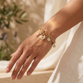 Close-up of a woman's wrist wearing a L'azurde gold charm bracelet (8.60g), illuminated by warm summer sunlight against soft ivory linen. لقطة قريبة لمعصم امرأة ترتدي سوار تعليقات ذهبي من لازوردي (8.60 غرام)، تضيئها أشعة شمس الصيف الدافئة على خلفية من الكتان العاجي الناعم.