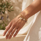 Close-up of a woman's wrist wearing a L'azurde gold charm bracelet (8.60g), illuminated by warm summer sunlight against soft ivory linen. لقطة قريبة لمعصم امرأة ترتدي سوار تعليقات ذهبي من لازوردي (8.60 غرام)، تضيئها أشعة شمس الصيف الدافئة على خلفية من الكتان العاجي الناعم.