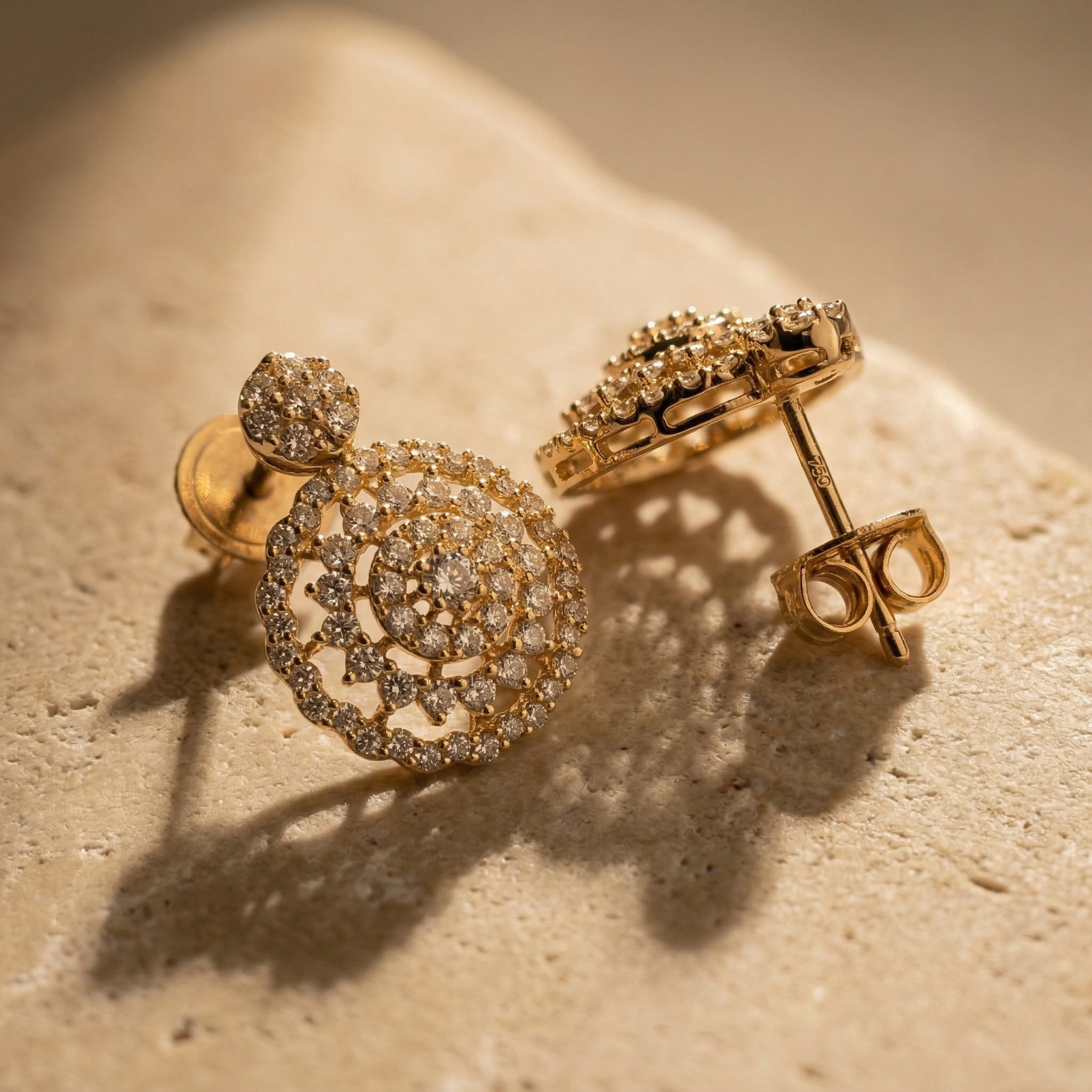A luxury close-up of Lazurde 18k yellow gold earrings resting on a textured, neutral stone surface, featuring soft, artistic shadows that highlight the pristine gold craftsmanship and glittering stone details.