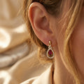 Close-up side profile of a woman wearing Lazurde 18k yellow gold drop earrings with a crimson center stone and diamond accents, set against a warm, elegantly blurred background.