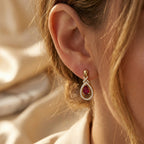 Close-up side profile of a woman wearing Lazurde 18k yellow gold drop earrings with a crimson center stone and diamond accents, set against a warm, elegantly blurred background.