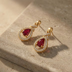 A pair of Lazurde 18k yellow gold earrings with crimson center stones resting beautifully on a textured neutral stone surface, styled with warm lighting and soft artistic shadows.