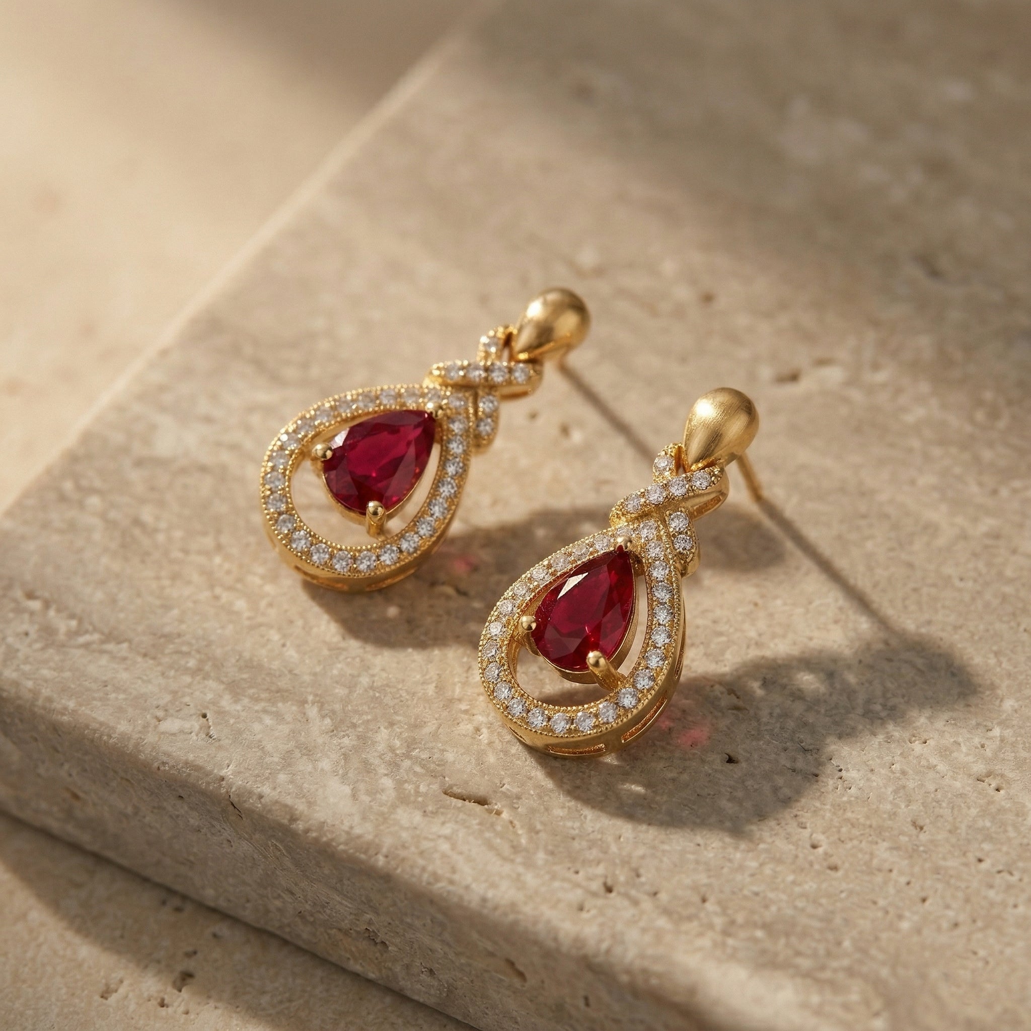 A pair of Lazurde 18k yellow gold earrings with crimson center stones resting beautifully on a textured neutral stone surface, styled with warm lighting and soft artistic shadows.