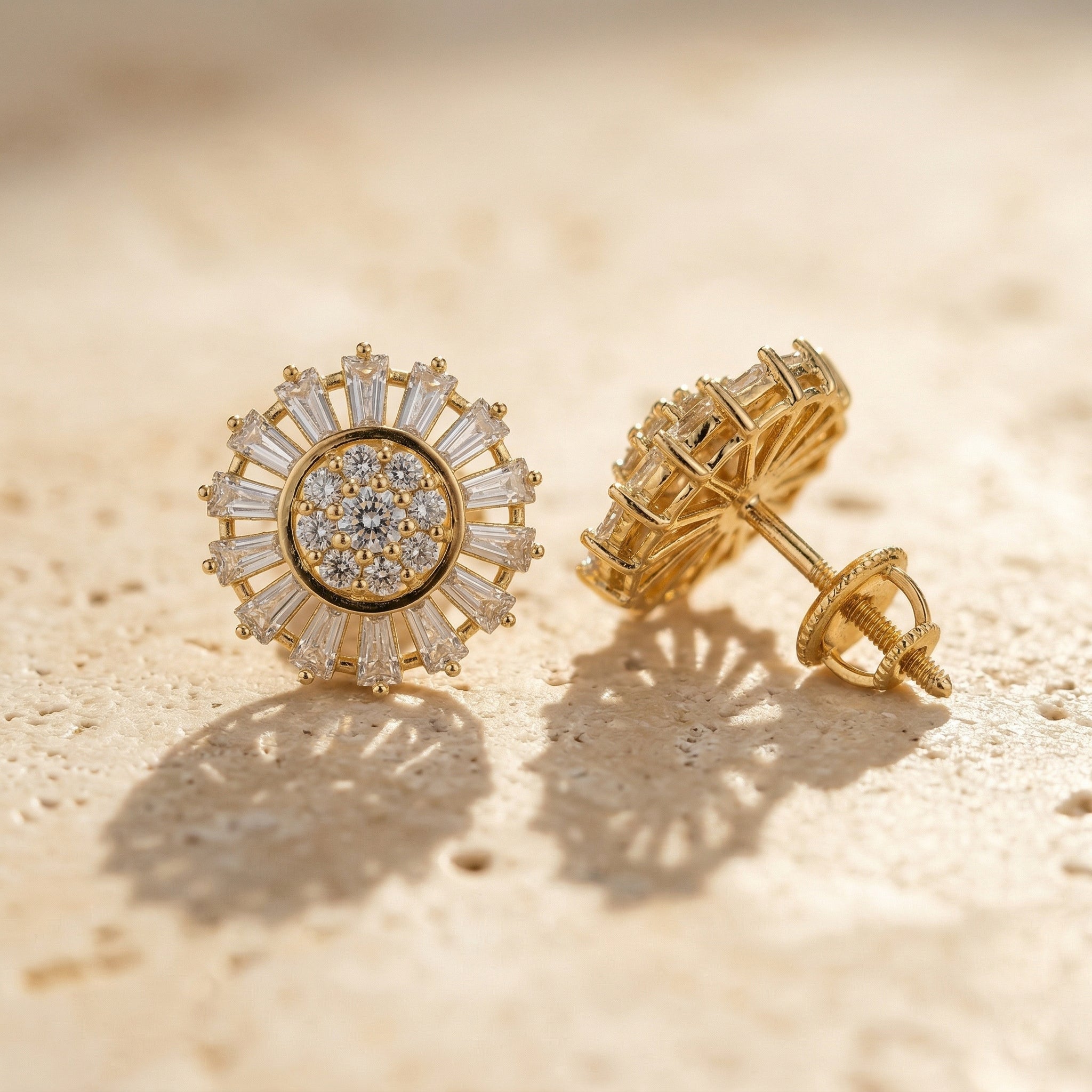 A high-end close-up of Lazurde 18k yellow gold earrings resting on a neutral, textured stone surface. Warm sunlight casts soft, artistic shadows that emphasize the meticulous craftsmanship and striking baguette-cut halo.