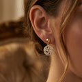 Close-up profile of a woman's ear modeling a Lazurde 18k yellow gold geometric drop earring. The jewelry is in sharp focus, showcasing the reflective baguette stones and warm gold tones against a blurred, elegant background with soft, warm lighting.