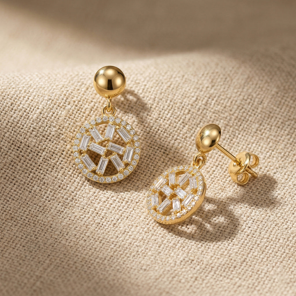 A pair of Lazurde 18k yellow gold drop earrings resting on a textured, neutral woven fabric. Warm, directional sunlight casts soft, artistic shadows, highlighting the high-polish gold craftsmanship and the brilliant details of the circular geometric pendants.