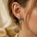 Side profile of a woman wearing Lazurde 18k yellow gold spike earrings with a trillion-cut teal gemstone, captured in soft warm lighting with a shallow depth of field for a luxury look.
