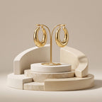 A high-end product display of a pair of chunky gold hoop earrings on a delicate gold stand. The stand sits on a tiered architectural podium made of curved cream and beige marble and stone elements, against a plain beige background.