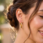 Elegant smiling woman outdoors wearing only 18k yellow gold elongated oval-grooved hoop earrings.