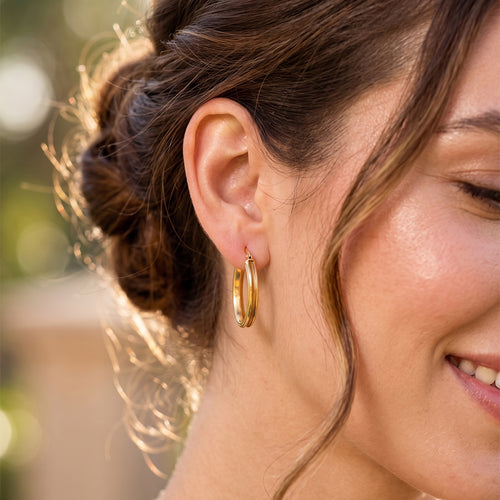 Elegant smiling woman outdoors wearing only 18k yellow gold elongated oval-grooved hoop earrings.