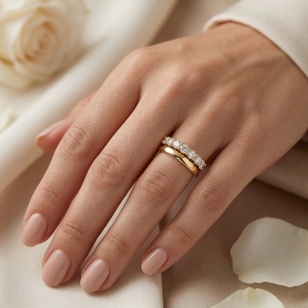 A close-up photograph of a woman’s left hand wearing two rings on her ring finger, showcasing the "Fine Gold" collection from Remas. A classic 18k yellow gold wedding band is paired with a matching 18k yellow gold wedding promise ring, featuring a row of small, round-cut diamonds. The hand is gently posed against a soft, ivory fabric background with a partially visible rose and petals, creating a delicate, bridal aesthetic.