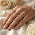A delicate Fine Gold 18k yellow gold wedding promise ring from The Forever Edit category, gracefully worn on a woman's hand and stacked with a classic gold band, resting against luxurious silk, lace, and soft cream roses.