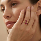 Close-up of a woman's hand wearing a gold ring with red gemstones, touching her face.
