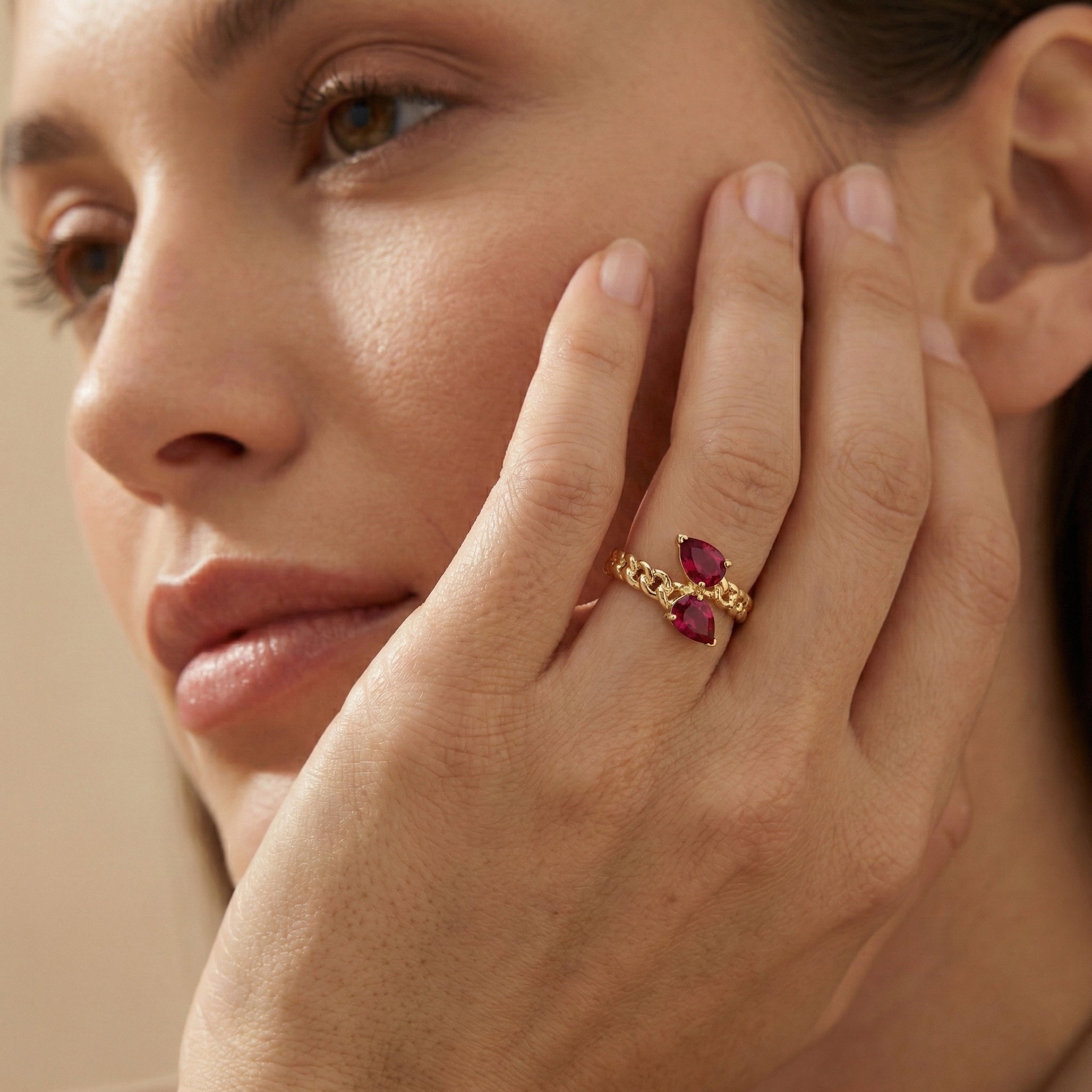 Close-up of a woman's hand wearing a gold ring with red gemstones, touching her face.