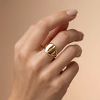 A luxury lifestyle photograph featuring an Andria 18k yellow gold ring worn elegantly on a feminine ring finger. The soft beige background and diffused lighting highlight the ring's sophisticated, bold silhouette.