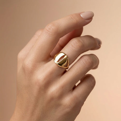 A luxury lifestyle photograph featuring an Andria 18k yellow gold ring worn elegantly on a feminine ring finger. The soft beige background and diffused lighting highlight the ring's sophisticated, bold silhouette.
