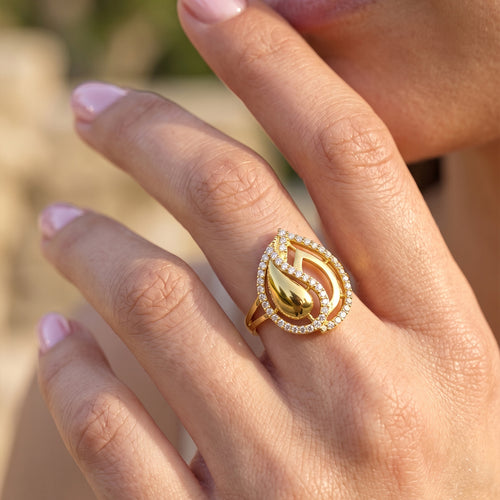 Close-up of a feminine hand gracefully modeling a Fine Gold 18k yellow gold ring with sparkling stones, set against a creamy, dreamy bokeh background. لقطة قريبة ليد أنثوية ترتدي خاتم ذهب عيار 18 من فاين جولد يبرز دقة التفاصيل ولمعان الفصوص، مع خلفية ناعمة وضبابية.