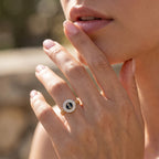 Close-up macro shot of a feminine hand gracefully modeling a Loaloaa 18k yellow gold black enamel initial ring against a dreamy outdoor bokeh background. لقطة قريبة ليد أنثوية ترتدي خاتم جنزير ذهب عيار 18 من لؤلؤة بمينا سوداء وتفاصيل دقيقة، مع خلفية خارجية ناعمة.