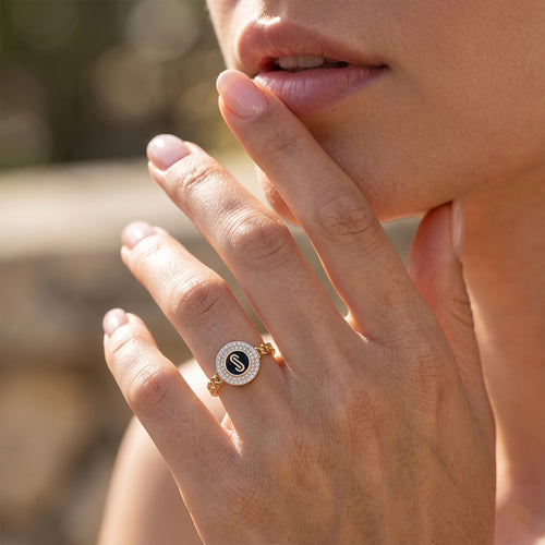 Close-up macro shot of a feminine hand gracefully modeling a Loaloaa 18k yellow gold black enamel initial ring against a dreamy outdoor bokeh background. لقطة قريبة ليد أنثوية ترتدي خاتم جنزير ذهب عيار 18 من لؤلؤة بمينا سوداء وتفاصيل دقيقة، مع خلفية خارجية ناعمة.