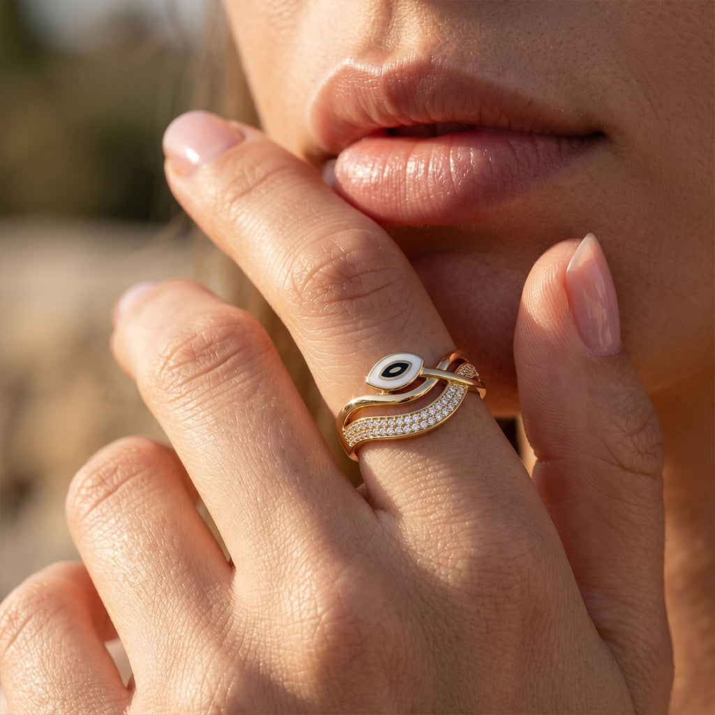 Close-up macro photo of a graceful feminine hand wearing a Dreams Gold 18k yellow gold enamel evil eye ring, set against a dreamy, warm outdoor bokeh background. لقطة قريبة ليد أنثوية ترتدي خاتم ذهب عيار 18 من دريمز جولد بتصميم العين وتفاصيل الفصوص الدقيقة، مع خلفية خارجية ناعمة.
