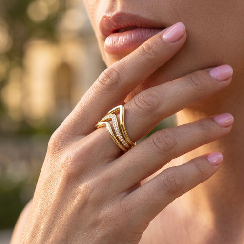 Close-up macro shot of the UNO 18k yellow gold V-shaped ring pointing elegantly downwards on a woman's hand, highlighting the sharp geometric design and sparkling center band.
صورة قريبة جداً (ماكرو) لخاتم أونو بتصميم السبعة مقلوب لتحت على إيد موديل، بتبرز شكل الـ V والفصوص اللي بتلمع في النص.