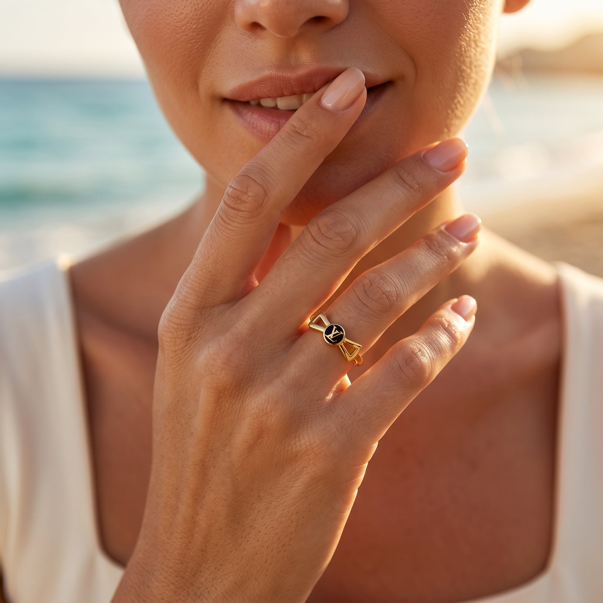 Delicate 18k yellow gold bow ring by Lamar worn on a woman's hand resting near her lips against a sunset beach backdrop.	خاتم رقيق من الذهب الأصفر عيار 18 من لامار ترتديه امرأة وتضع يدها بالقرب من شفتيها مع خلفية شاطئ وقت الغروب.